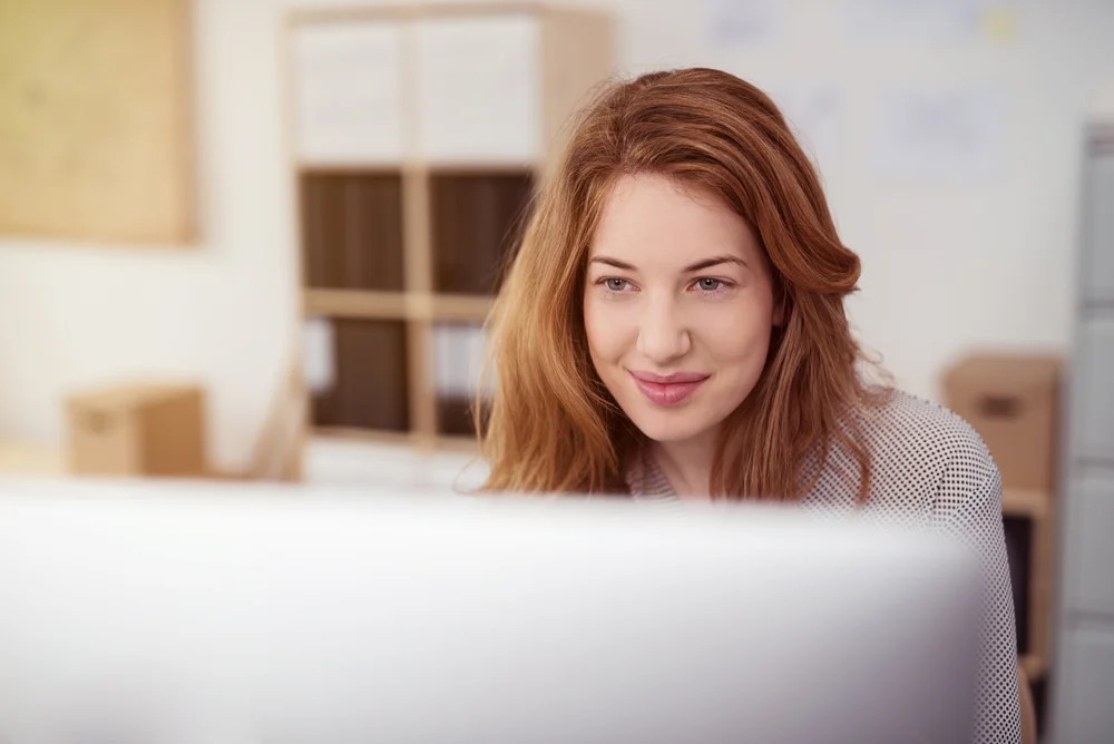 Attractive young woman working on a desktop computer smiling as she leans forwards reading text on the screen view over the monitor