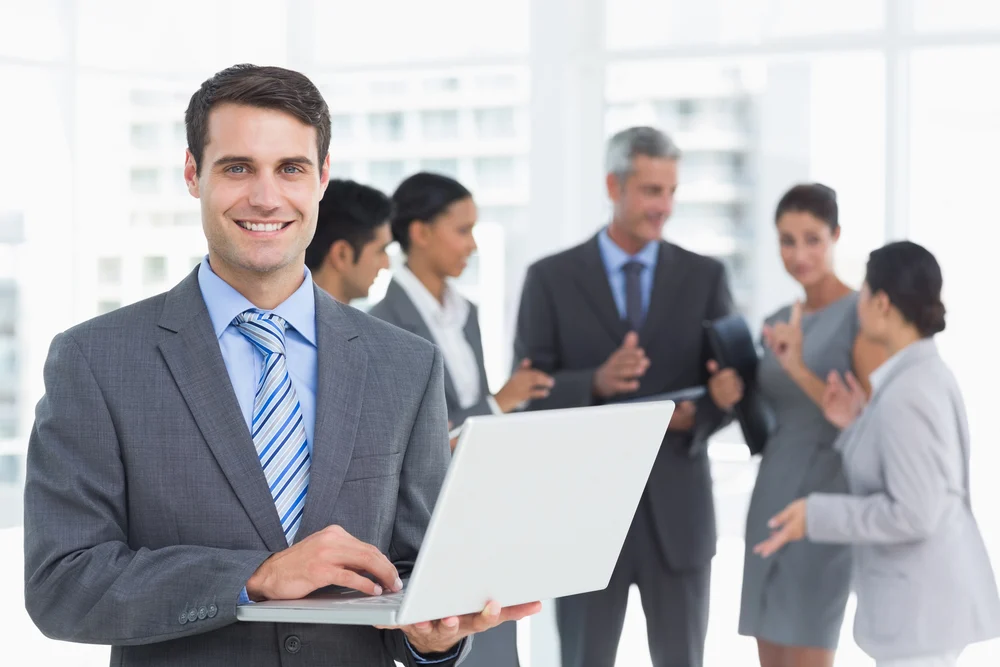 Businessman using laptop with colleagues behind in the office Businessman using laptop with colleagues behind in the office